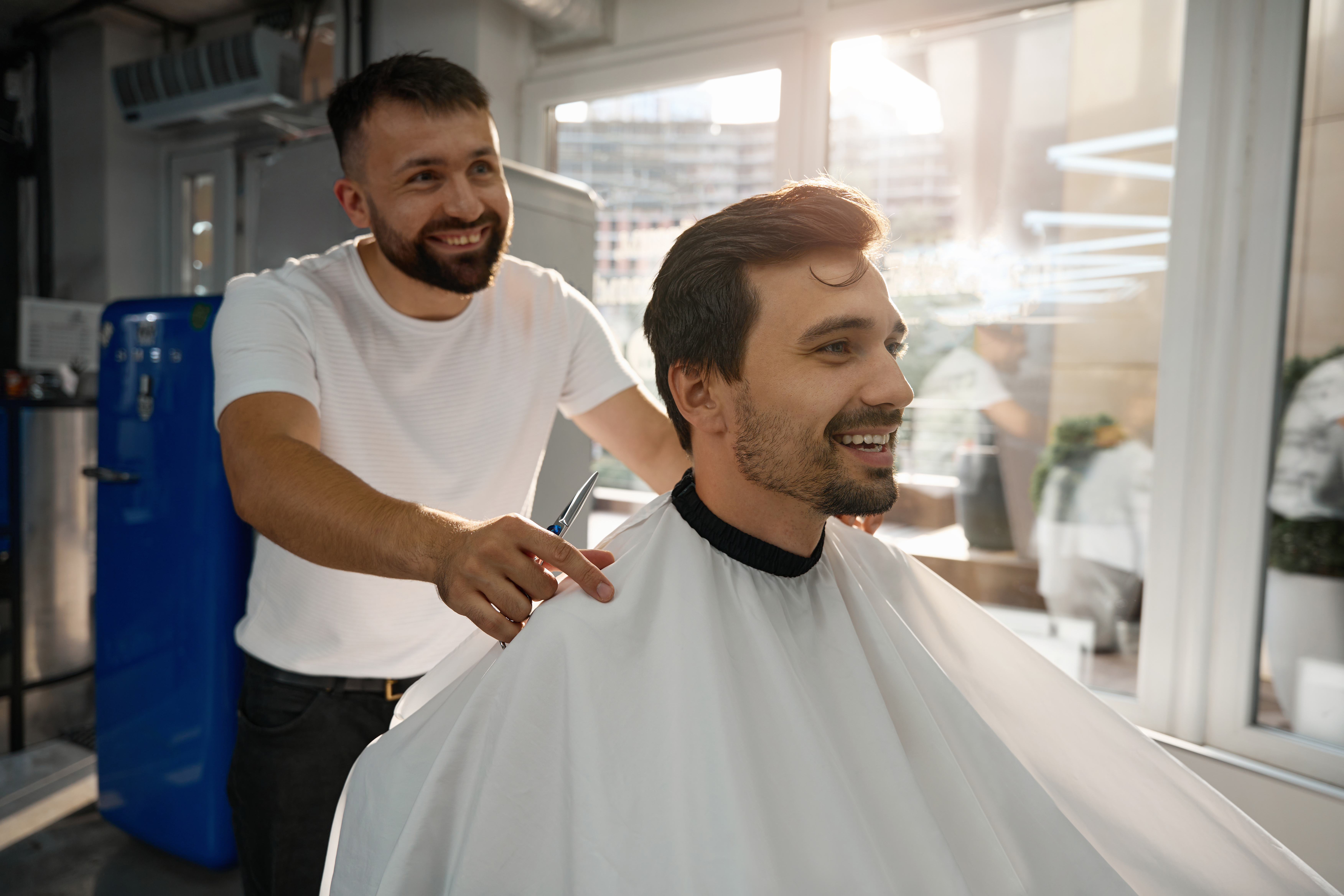 Contented brunette man and happy barbershop worker smiling while being satisfied with the results of hair-grooming